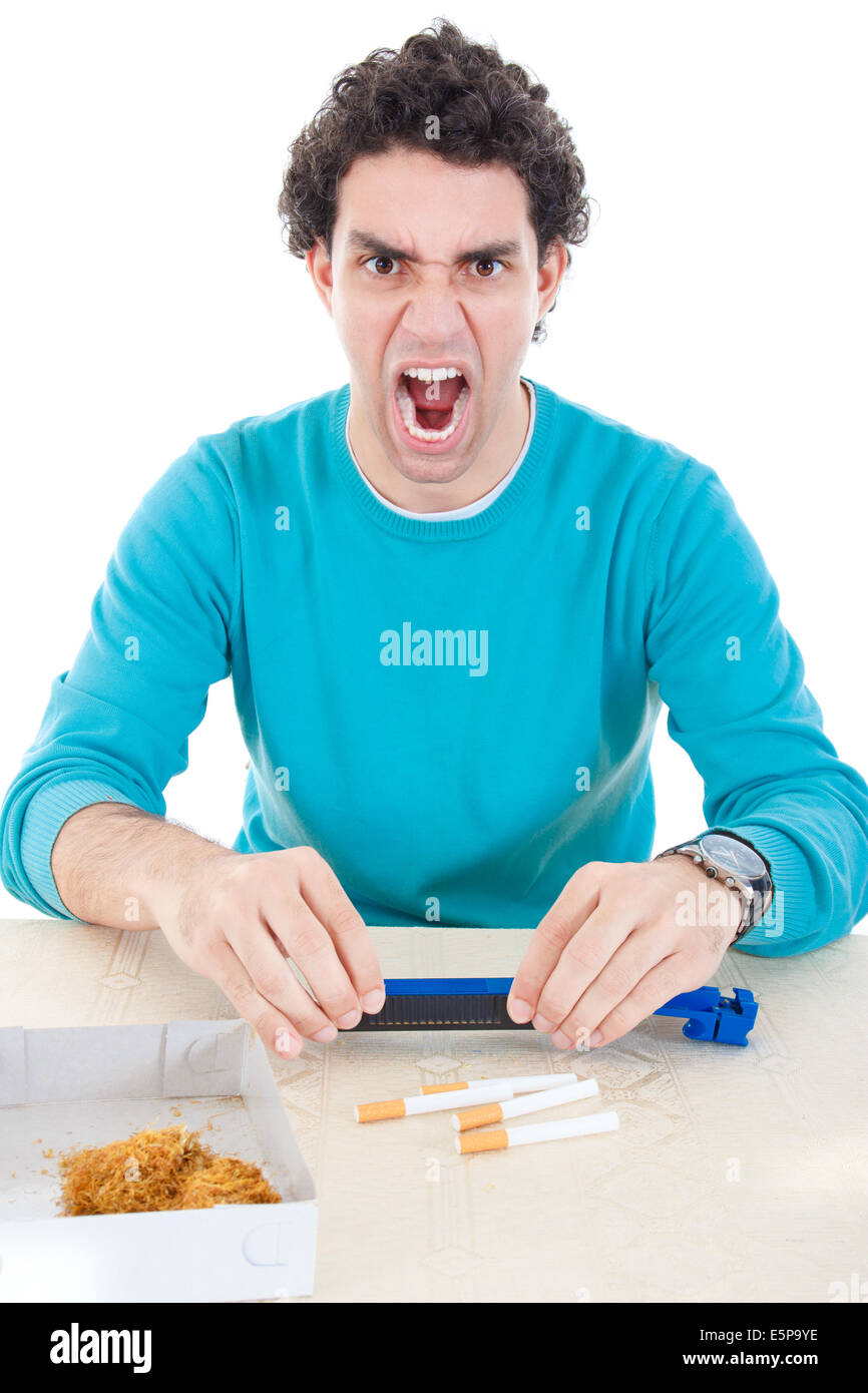 El hombre enojado en jersey azul haciendo cigarrillos con dispositivo de cigarros y tabaco seco sobre la mesa, la crisis económica por alto precio de CI Foto de stock