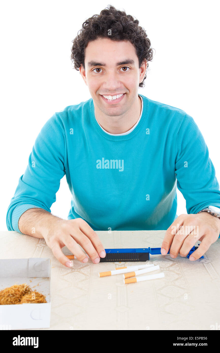 Hombre sonriente en jersey azul haciendo cigarrillos con dispositivo de cigarros y tabaco seco sobre la mesa Foto de stock