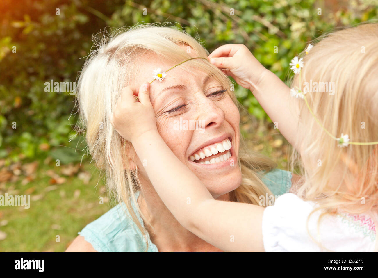 Cerca del infante femenino colocando daisy chain en el cabello de la madre en el jardín Foto de stock