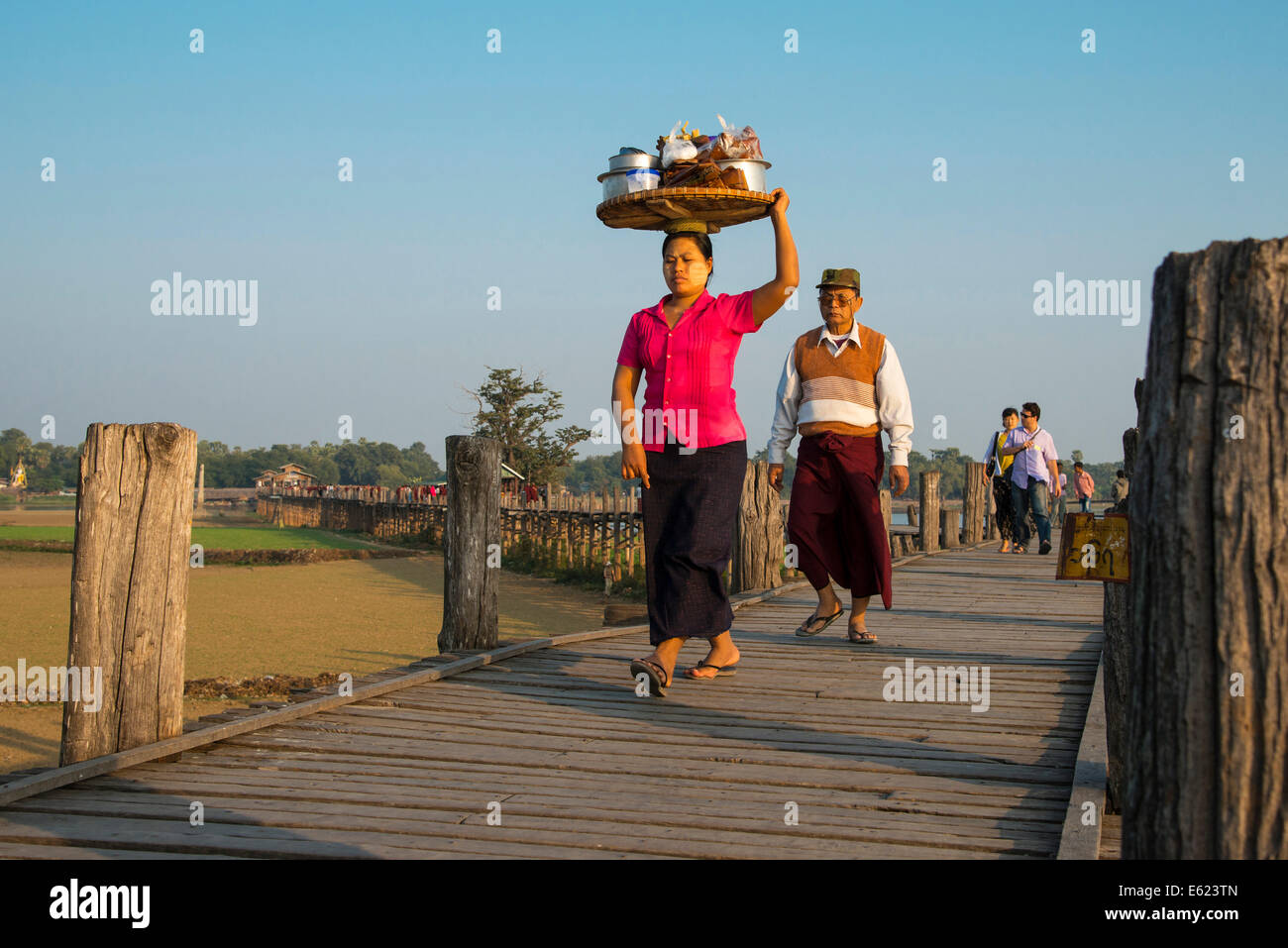 Mujer con tanaka en su cara llevar comida en su cabeza en un puente de teca, U Bein Puente sobre el lago, Amarapura Thaungthaman Foto de stock