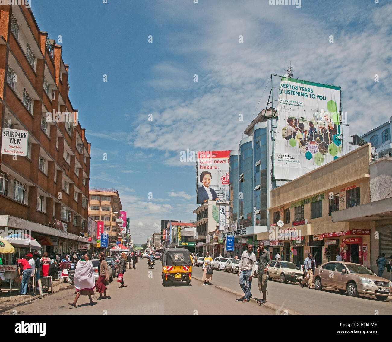 La gente y el tráfico en la avenida Kenyatta Nakuru, Kenya, Africa oriental con vallas publicitarias tiendas y tres ruedas Piaggio Ape taxi Foto de stock