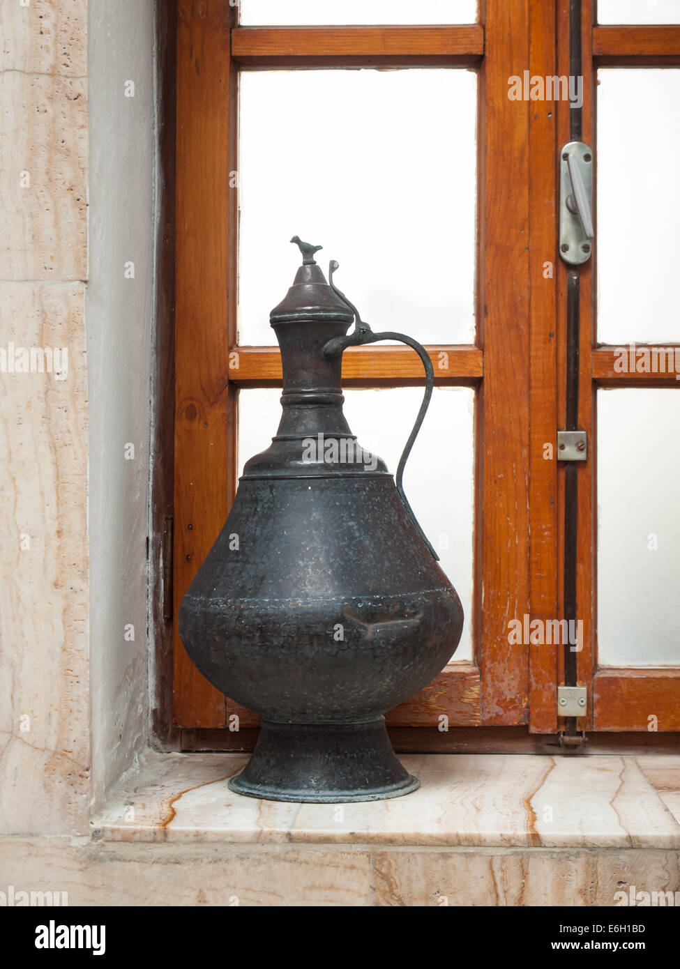 Metal antiguo jarra de agua sobre un alféizar de baño en mármol marrón con ventana escarchada detrás Foto de stock