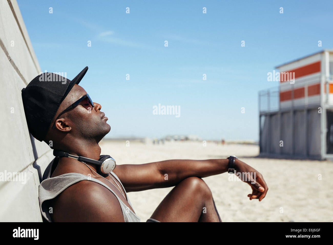 Vista lateral del hombre africano relajado a una pared inclinada en la playa tomando el sol. Chico negro con gafas de sol y relax cap. Foto de stock
