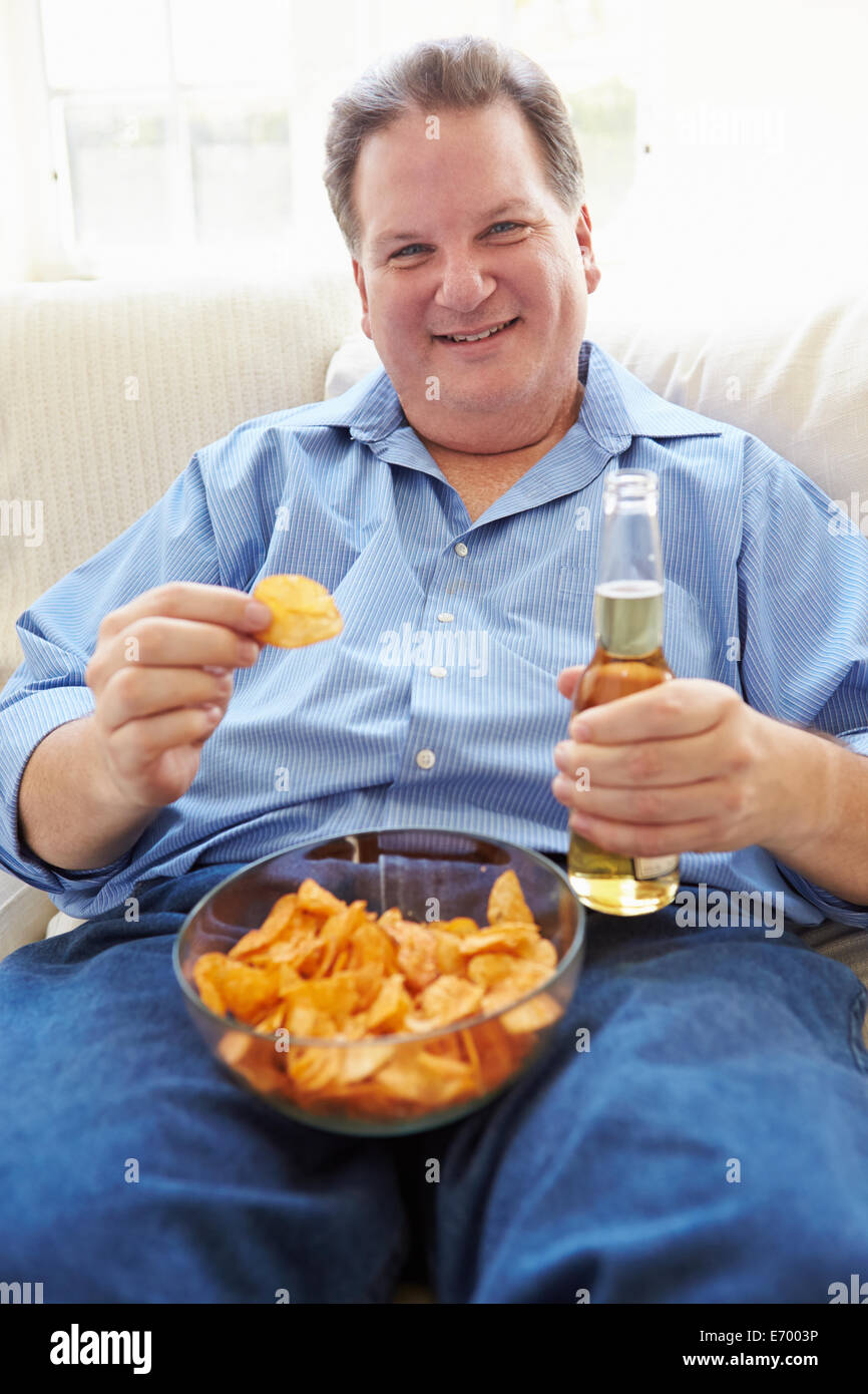 Sobrepeso hombre en casa comiendo papas fritas y bebiendo cerveza Foto de stock