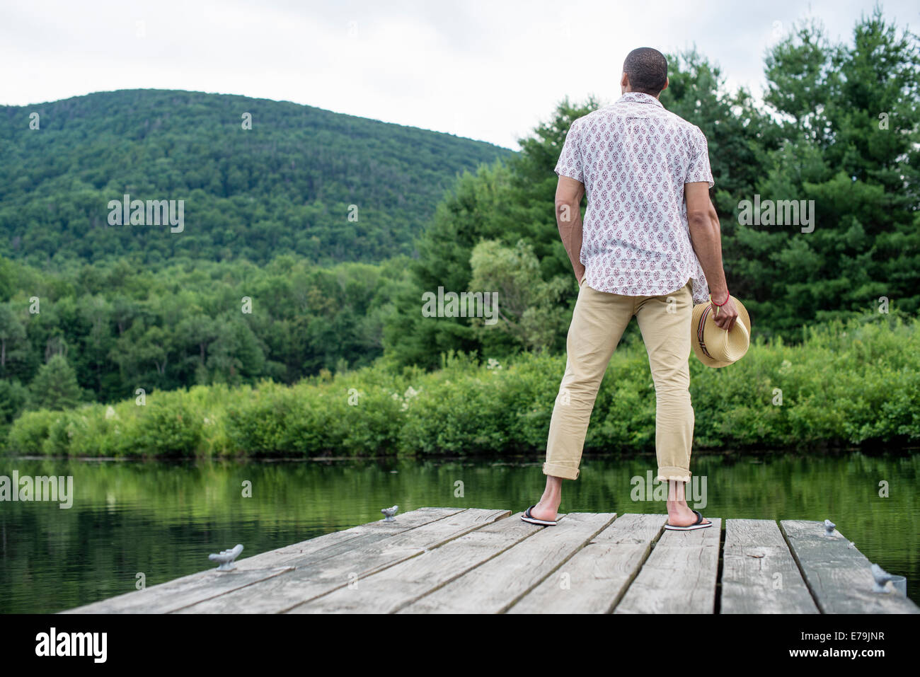 Un hombre de pie en un muelle de madera con vistas a un tranquilo lago. Foto de stock