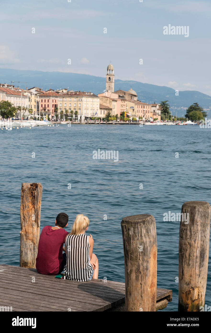 Pareja joven sentarse y disfrutar de las vistas y del sol de la tarde en la orilla del lago de Garda en Salo en el lado este del lago Foto de stock