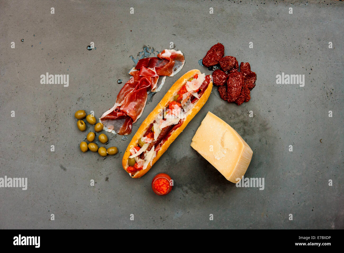 Moderno hot dog con salchicha de cordero, jamón, tomates secados al sol, queso parmesano, aceitunas, salsa de tomate de mesa de hormigón Foto de stock