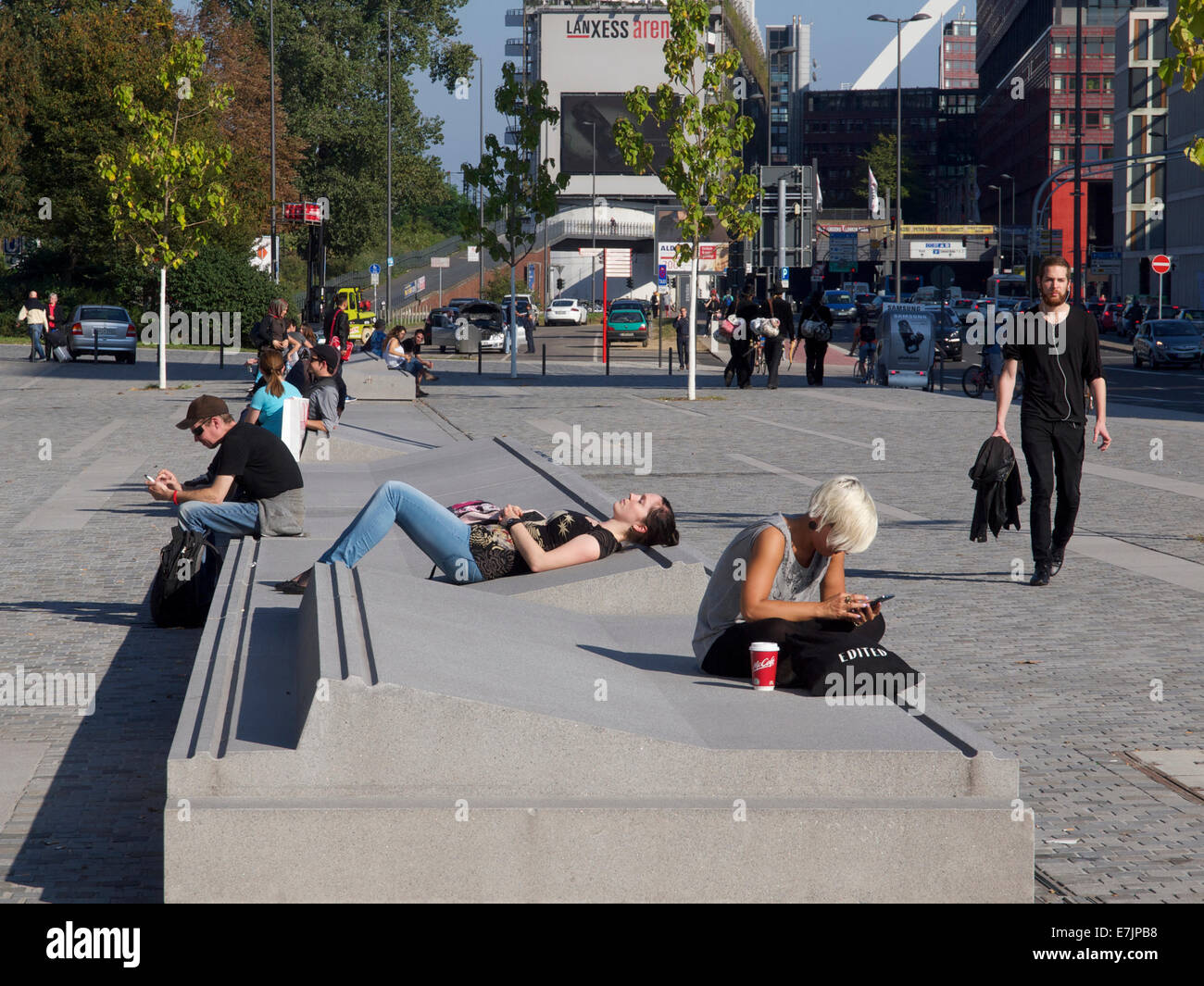 Mobiliario urbano de hormigón con jóvenes relajándose en el sol y el uso de teléfonos móviles en la estación de tren de Colonia, Alemania Foto de stock