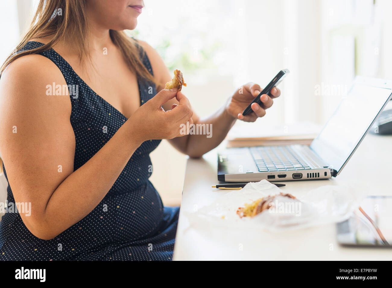 Mujer comiendo croissant y a través de teléfono móvil en la oficina Foto de stock