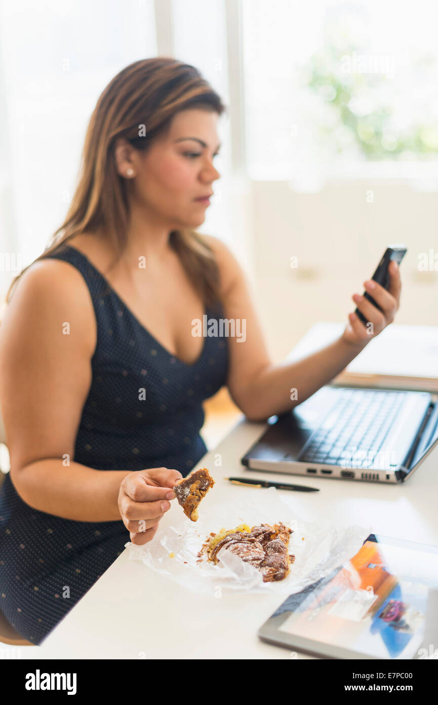 Mujer comiendo croissant y a través de teléfono móvil en la oficina Foto de stock