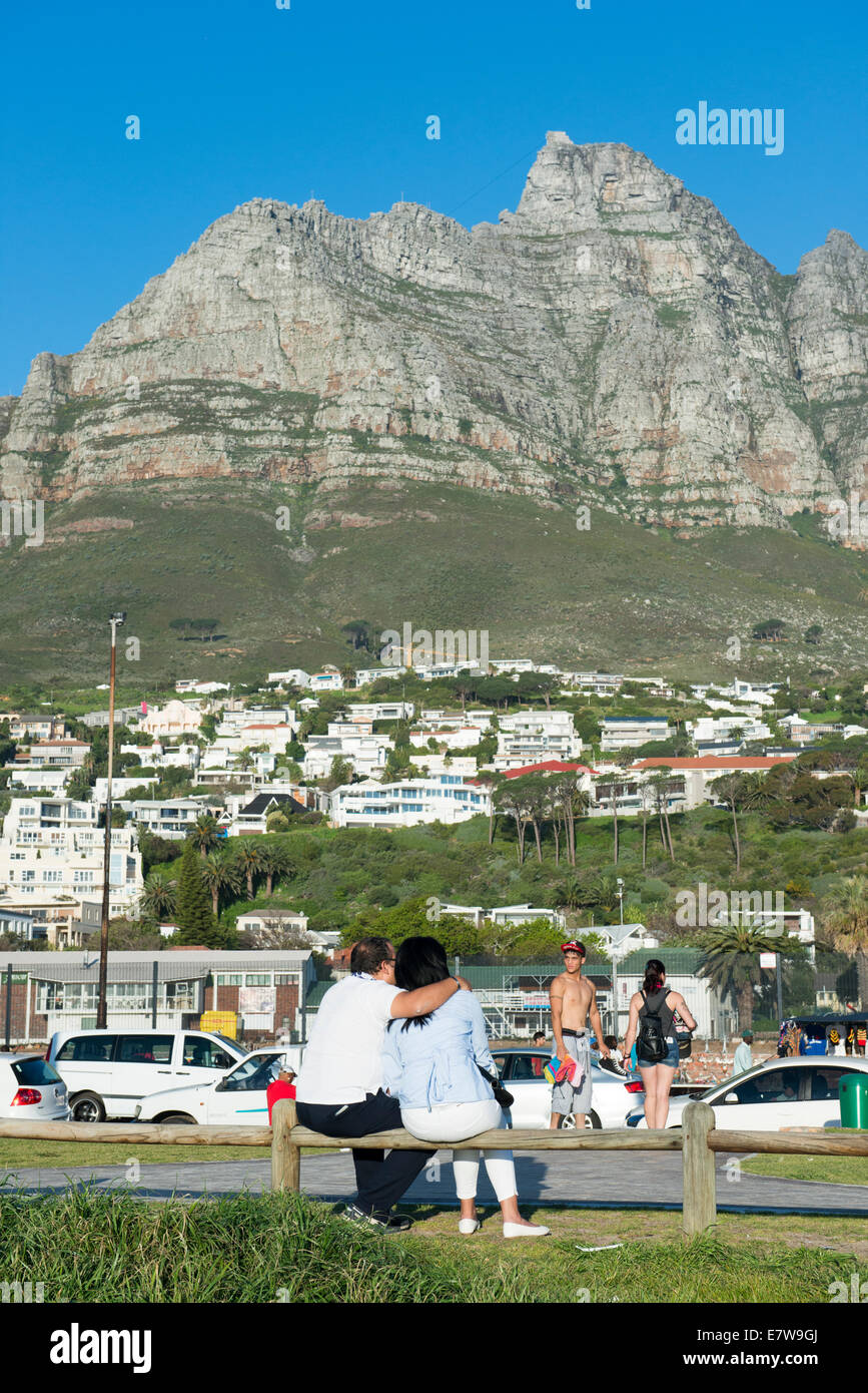 Una pareja sentada en un banco en Camps Bay, la montaña de la Mesa en el fondo, Ciudad del Cabo, Sudáfrica Foto de stock