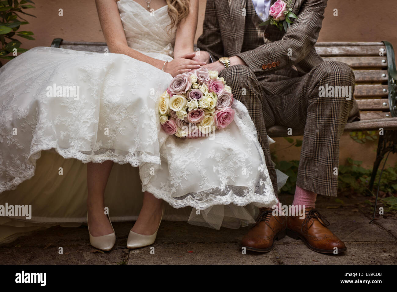 Un novios vistiendo un traje de tweed vintage style& 50's style boda,sentado en un banco, tomados de la mano y un ramo de rosas Foto de stock