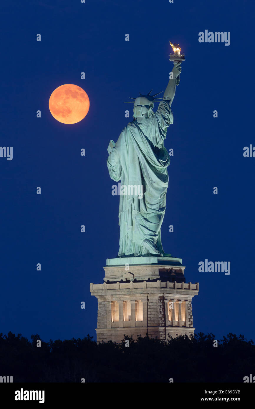 La super luna se eleva junto a la Estatua de la libertad durante la hora azul. Foto de stock