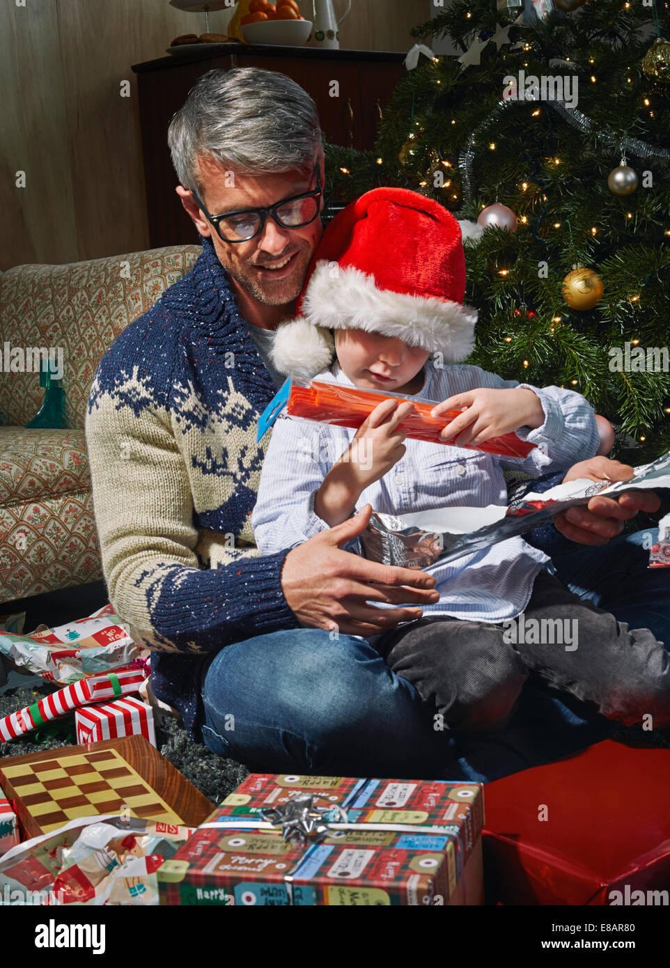 Padre en el suelo de la sala de estar abriendo regalos de navidad con hijo Foto de stock