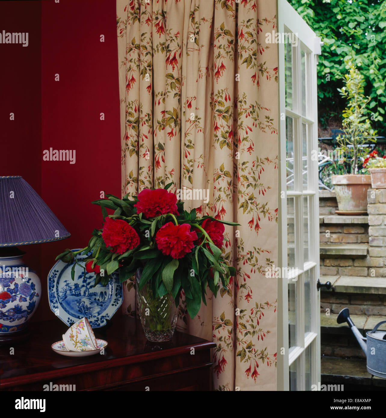 Las Peonías rojas en un jarrón sobre la mesa junto a la ventana francesa con cortinas con motivos florales en el país comedor Foto de stock