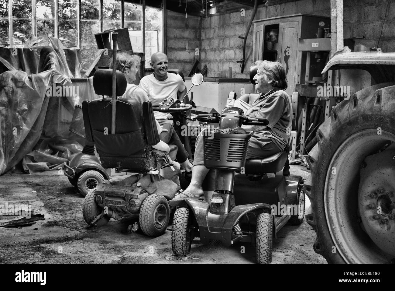 Tres personas sentadas sobre movilidad scooters chateando en un tractor viejo cobertizo Foto de stock
