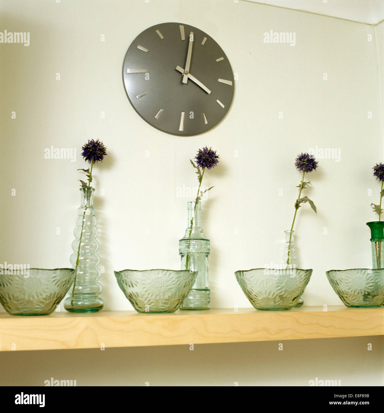 Reloj circular en pared sobre la estantería con recipientes de cristal texturizado y botellas con flores Foto de stock