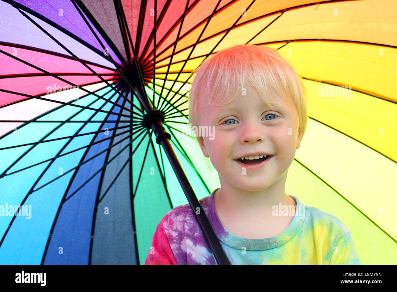 Un poco feliz de dos años, el niño está sonriendo como él está fuera, bajo un arco iris de colores paraguas para mantenerlo seco de la lluvia Foto de stock