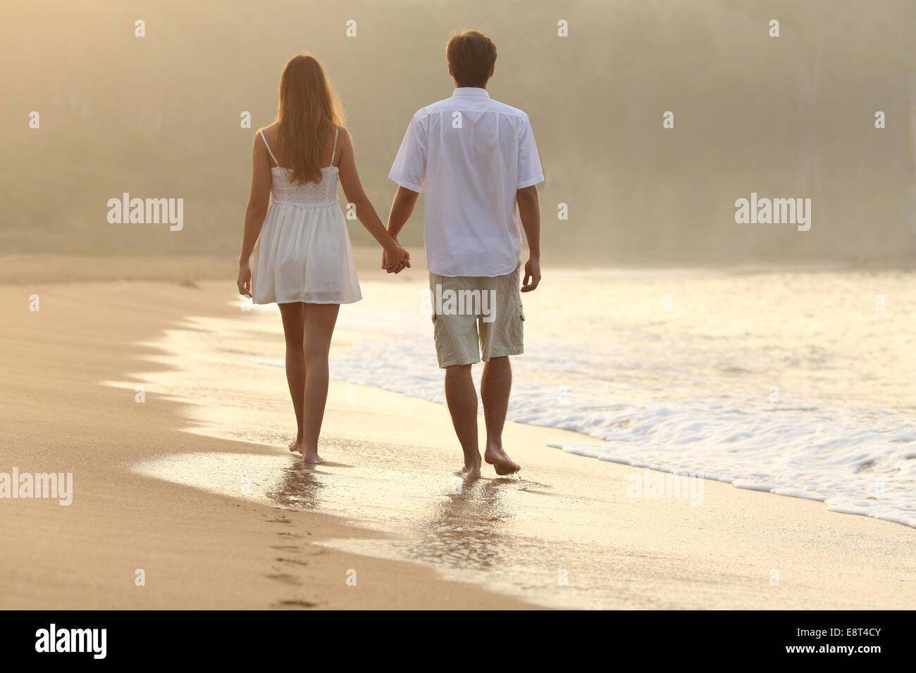 Vista posterior de una pareja caminando y tomados de la mano en la arena de una playa en el atardecer. Foto de stock