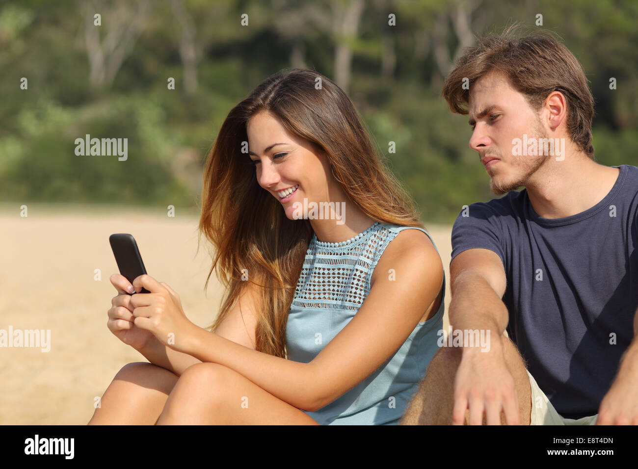 Novio celoso viendo a su novia mensajes de texto en el teléfono en la playa Foto de stock