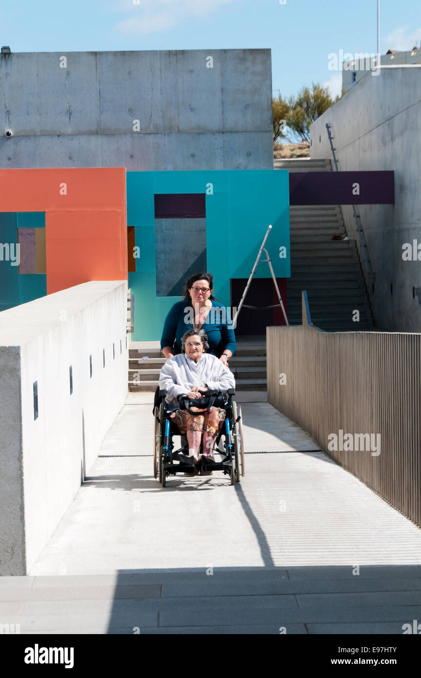 Una anciana es empujado hasta una rampa de acceso para discapacitados de hormigón en una silla de ruedas por su cuidador o auxiliar en un día soleado. Foto de stock