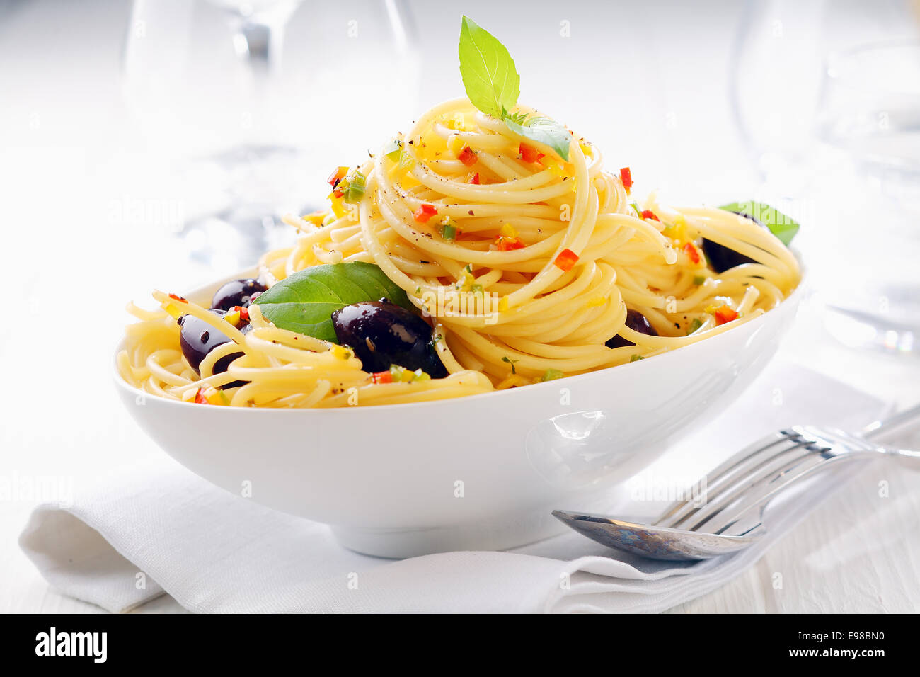 Primer plano de espaguetis pasta en un recipiente blanco con aceitunas en una servilleta blanca con copas de vino en el fondo. Foto de stock