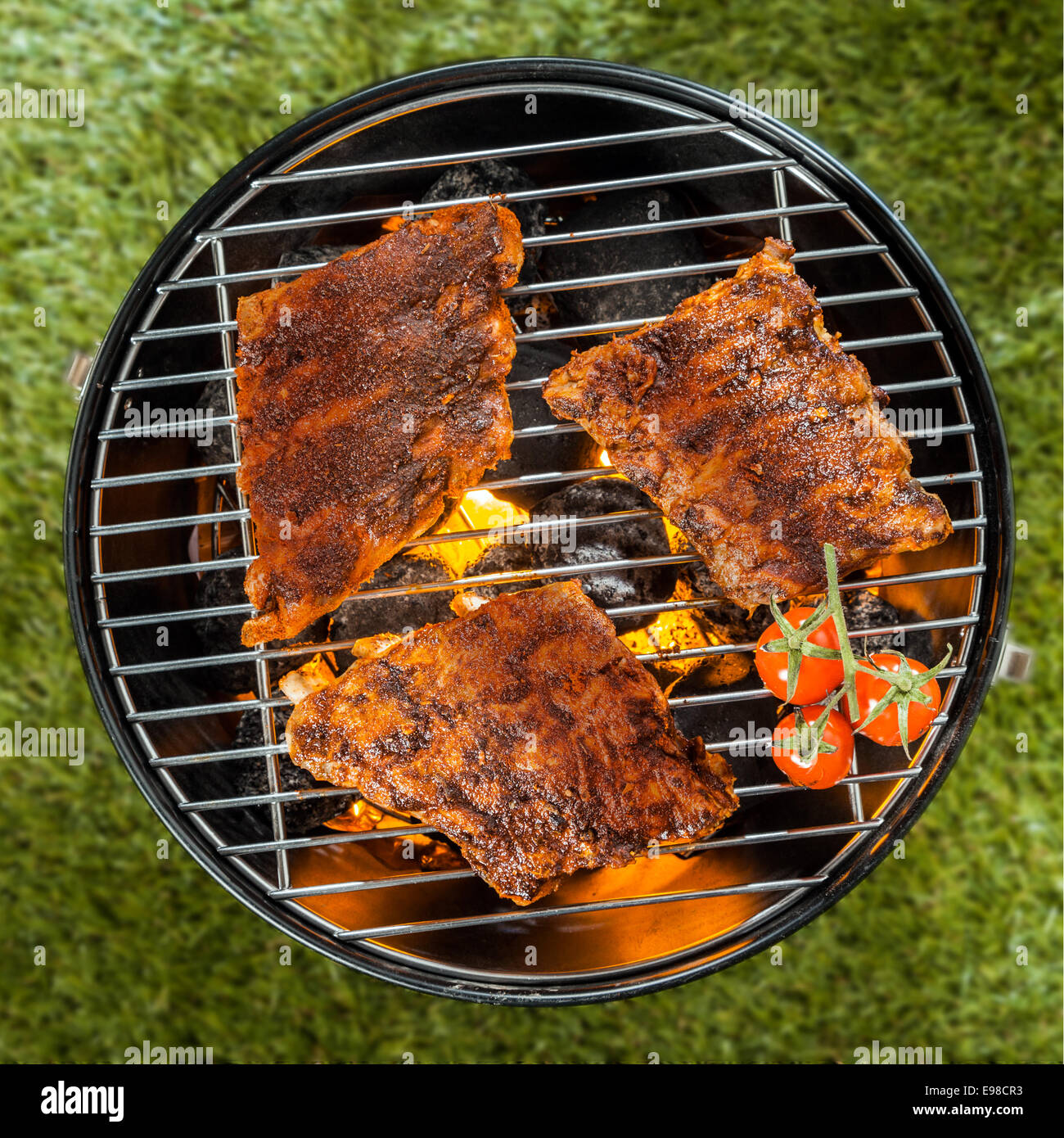 Vista desde arriba de tres deliciosas sazonado racks de costilla cocinar sobre un fuego en una barbacoa portátil de metal redondo barbacoa en el pasto verde Foto de stock