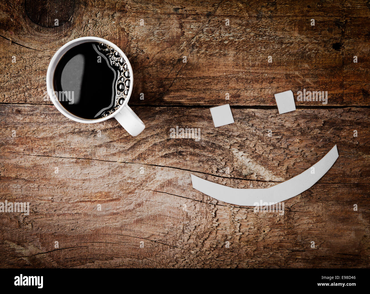 Vista aérea de una taza caliente de café espresso negro fuerte con una cara sonriente con ojos de cubos de azúcar sobre un fondo de madera con textura rugosa Foto de stock