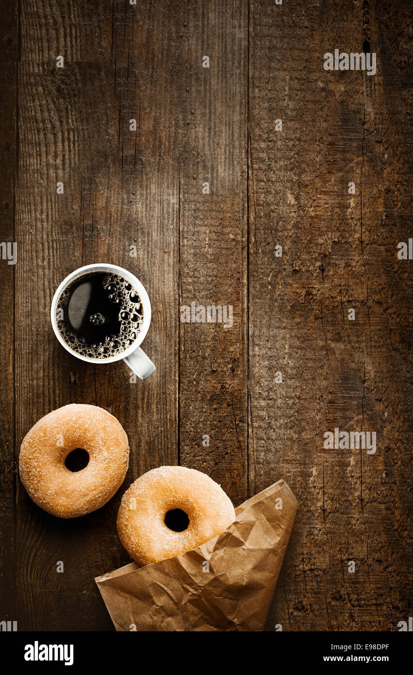 Vista aérea de dos tentador dulce donas azucaradas con su envoltorio de papel marrón y una taza de fuerte café espresso o filtro de negro sobre una tabla de madera rústica Foto de stock