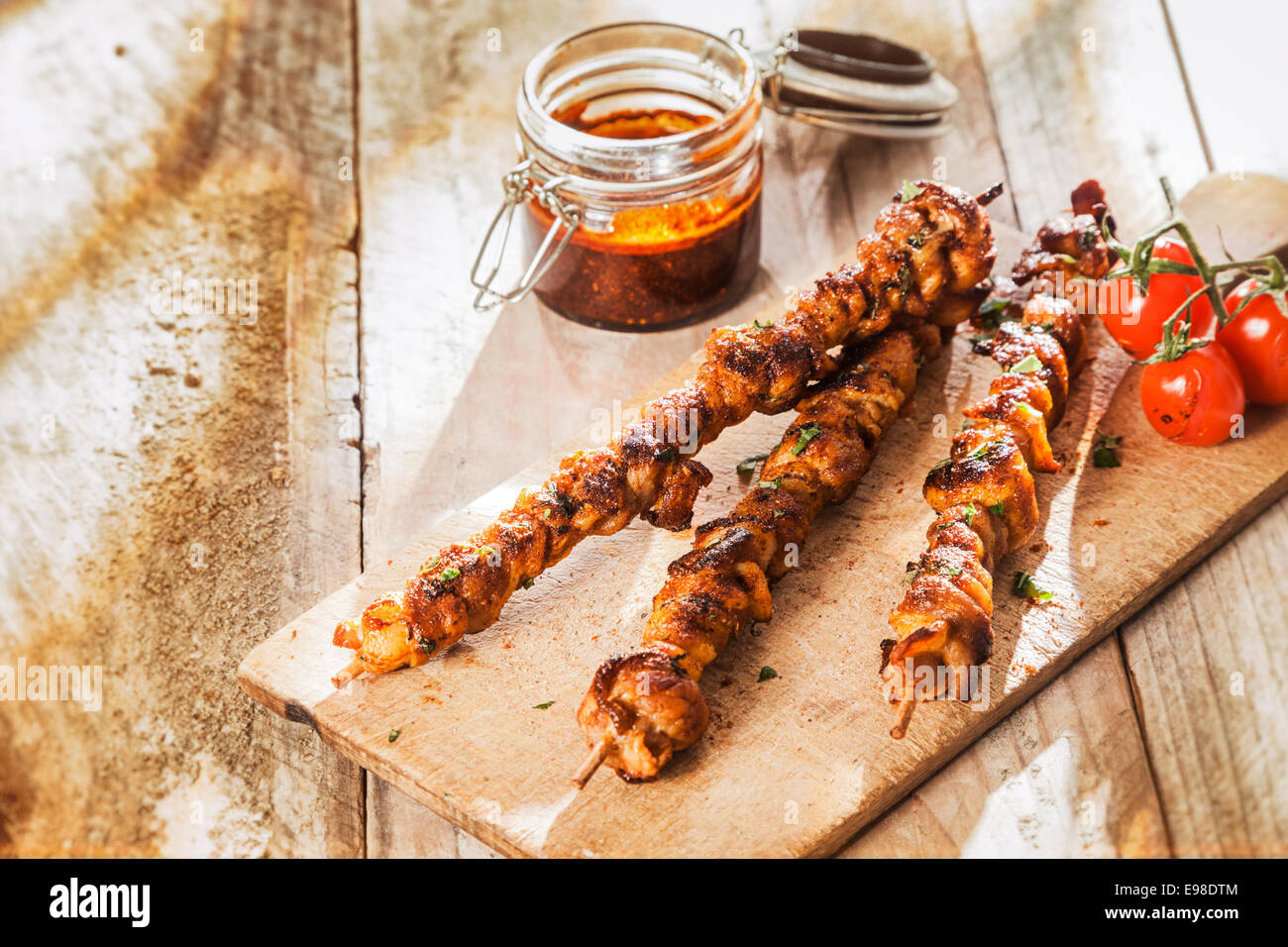 Las brochetas de carne a la parrilla picante en un picnic de verano servido sobre una tabla de cortar de madera con tomates asados junto a un frasco de picante salsa, rociándolo con copyspace Foto de stock