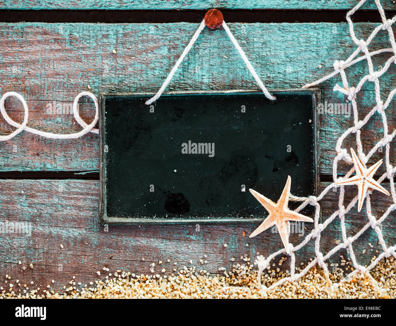 Todavía la vida marina con una pizarra en blanco letrero con copyspace para su texto, menú o anuncio decorado con red de pesca y Starfish sobre tablas de madera pintadas de azul sobre la arena del mar Foto de stock