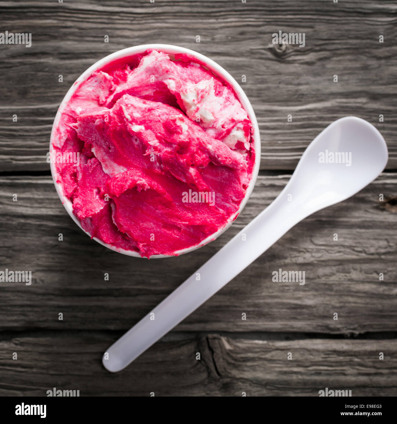 Delicioso helado de frambuesa refrescante fría en una tina de plástico para llevar con una cucharadita sobre una superficie de madera con textura antigua, sobrecarga Foto de stock