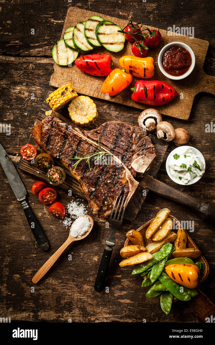 Propagación sana con t-bone o porterhouse steak servido con un surtido de verduras asadas sanos y sabrosos dips sobre una tabla de madera rústica en una cocina de campo Foto de stock