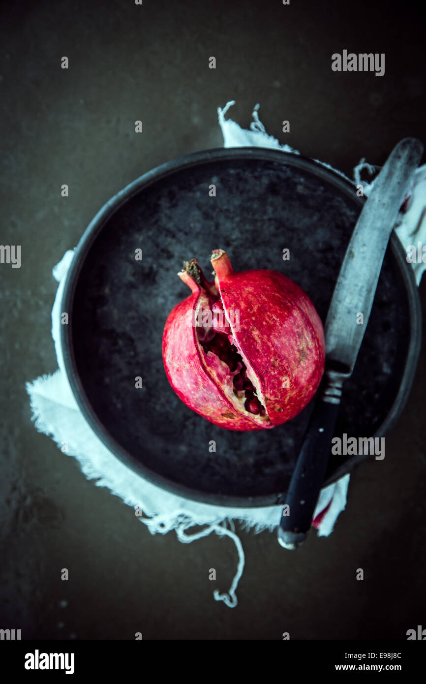 Dividir una granada abierta con un gran cuchillo de cocina para acceder a las jugosas semillas rojas maduras, vista desde arriba en una placa contra un fondo oscuro Foto de stock