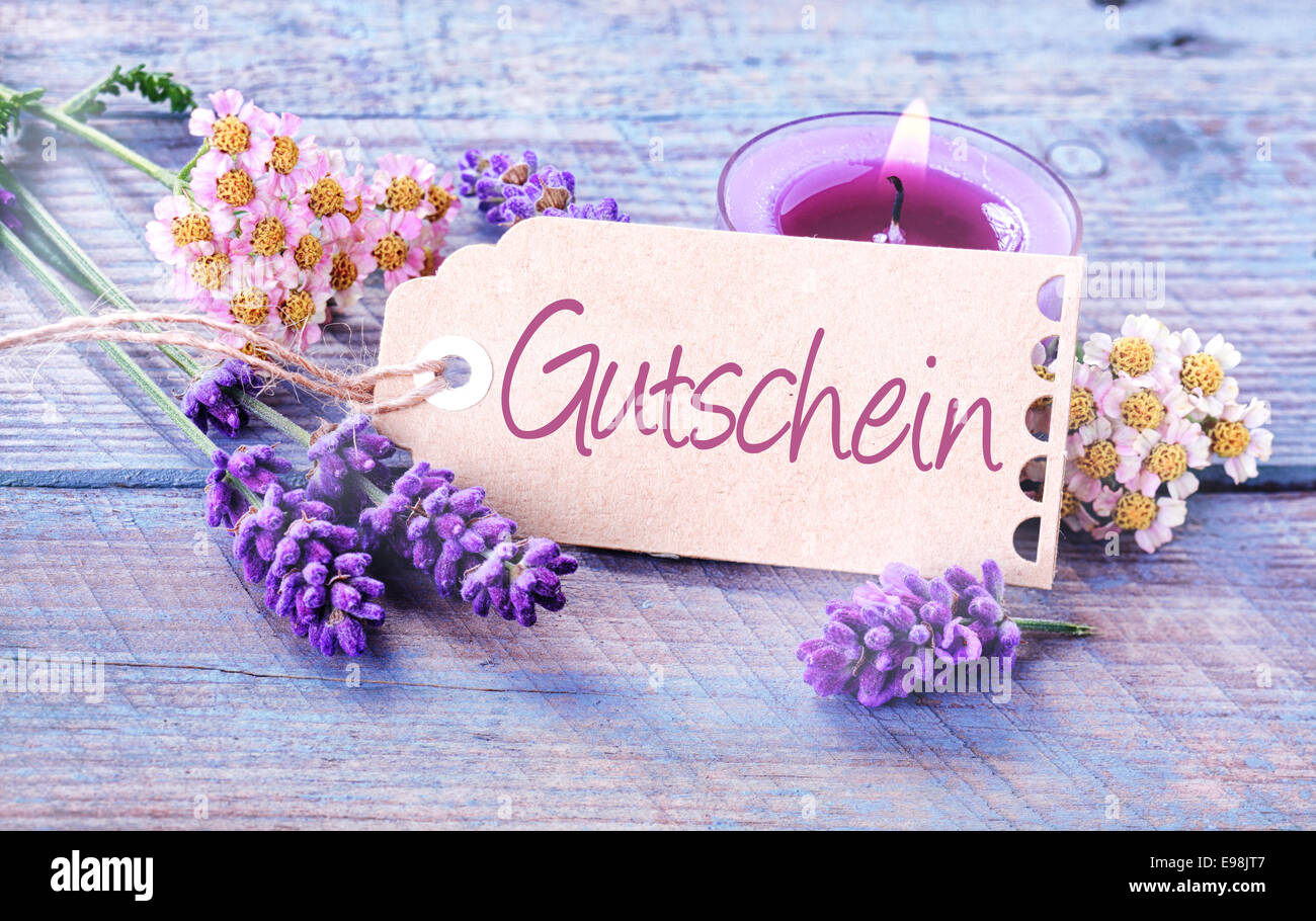 Etiqueta del regalo con la palabra Gutschein en alemán sobre tablones de madera azul claro con una vela encendida y perfumada, lavanda y flores frescas en un concepto de spa y bienestar Foto de stock