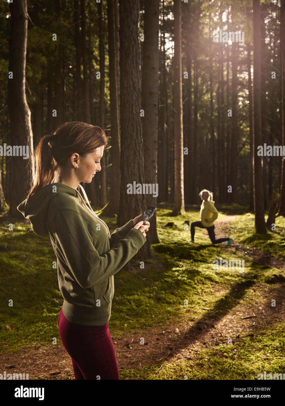 Mujer de pie en el bosque comprobando su smartphone, otra mujer haciendo ejercicios de estiramiento en la parte posterior, Tirol, Austria Foto de stock
