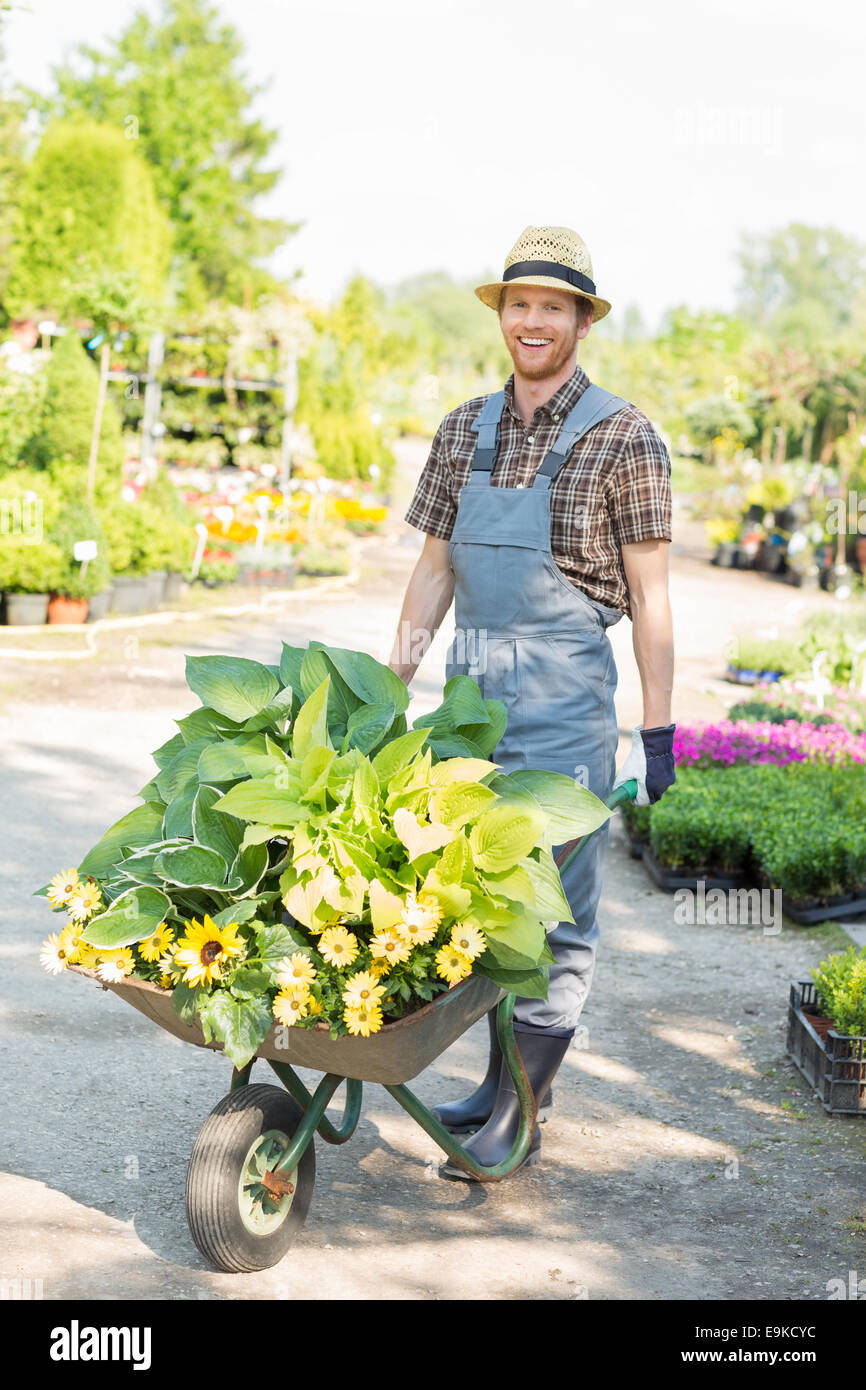Retrato de cuerpo entero del jardinero feliz empujando la carretilla de mano con las plantas en el jardín Foto de stock