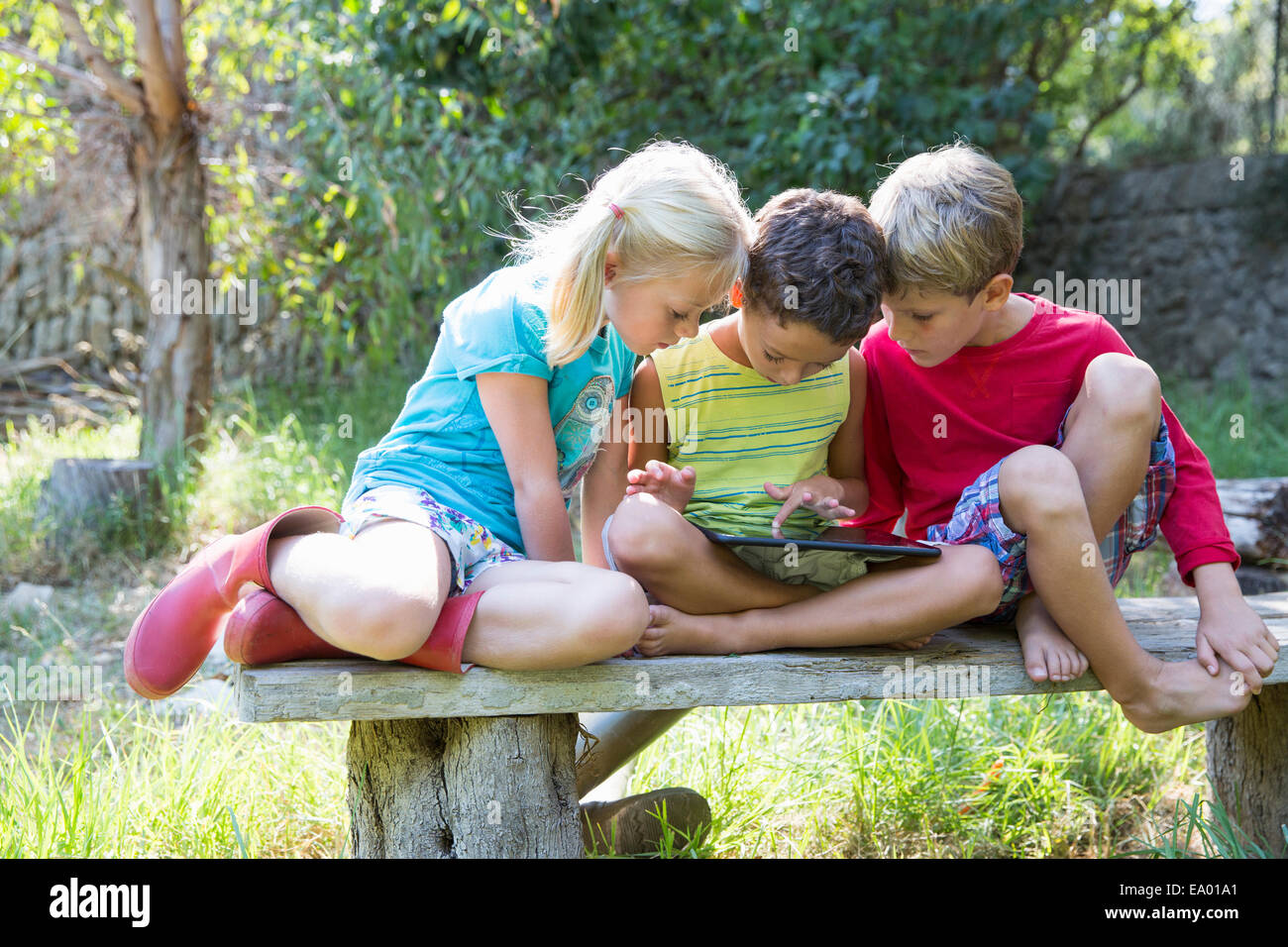Tres niños sentados en el asiento de jardín mirando hacia tableta digital Foto de stock