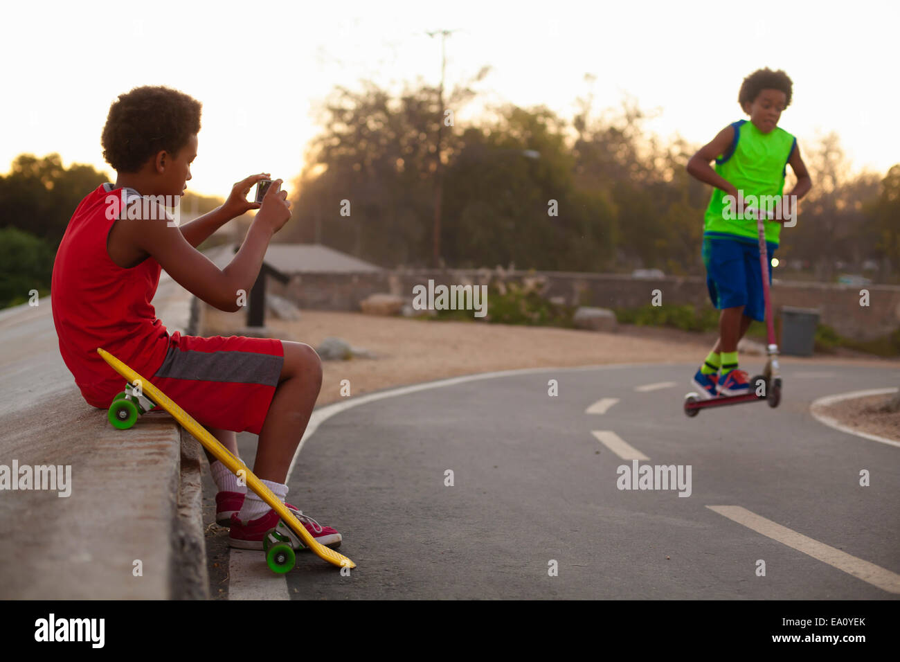 Boy fotografiar hermano haciendo push scooter saltar en carretera Foto de stock