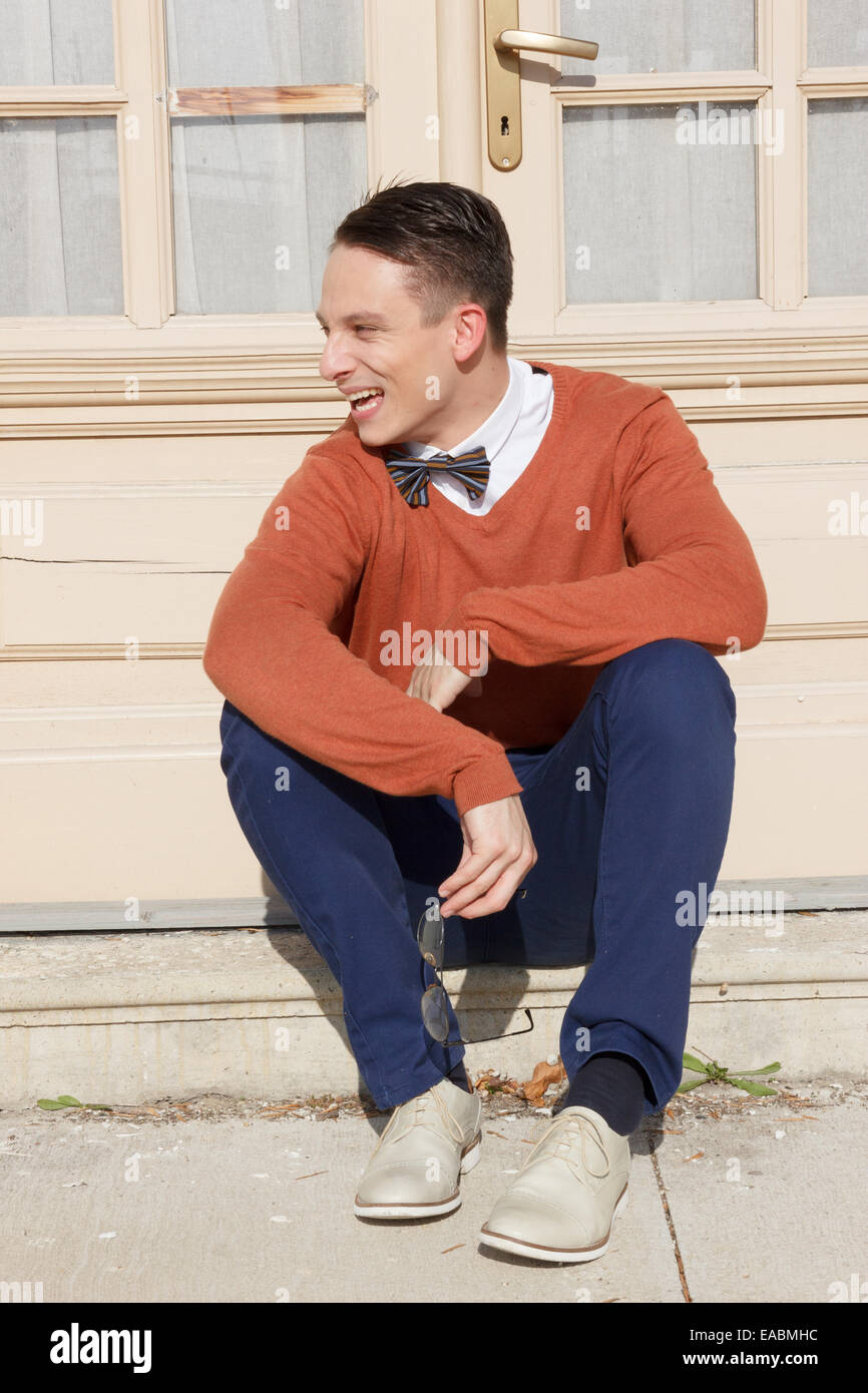Feliz joven de suéter sentado en pasos en la parte frontal de la casa y planteando , vintage retro foto Foto de stock