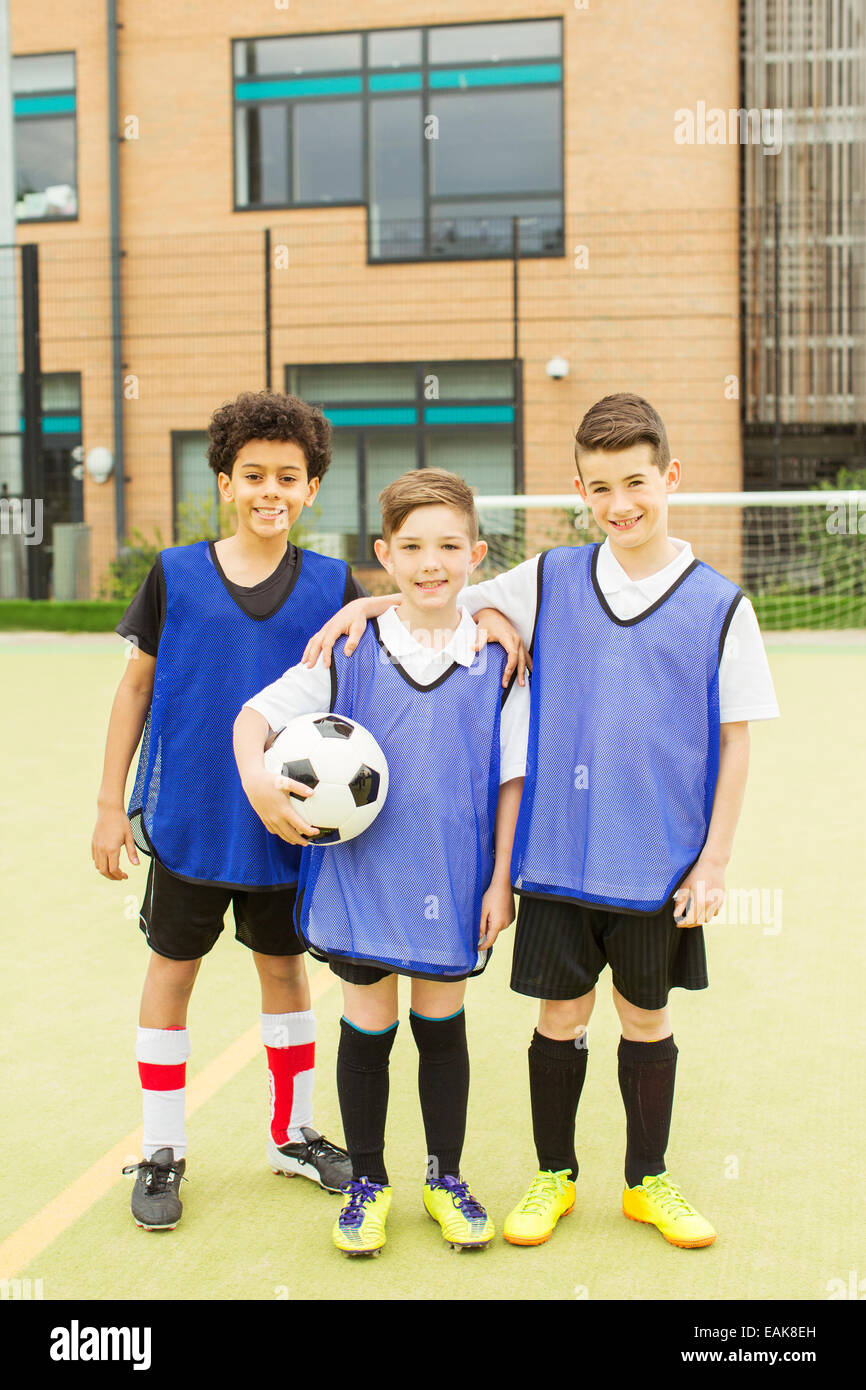 Retrato de tres niños sonrientes vistiendo uniformes de fútbol y sosteniendo un balón de fútbol delante de la escuela Foto de stock