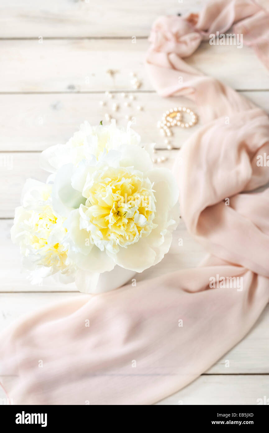 Un montón de peonías cremosa blanca en una jarra blanca sobre una tabla de madera blanca con una luz rosa bufanda y perlas en el fondo Foto de stock