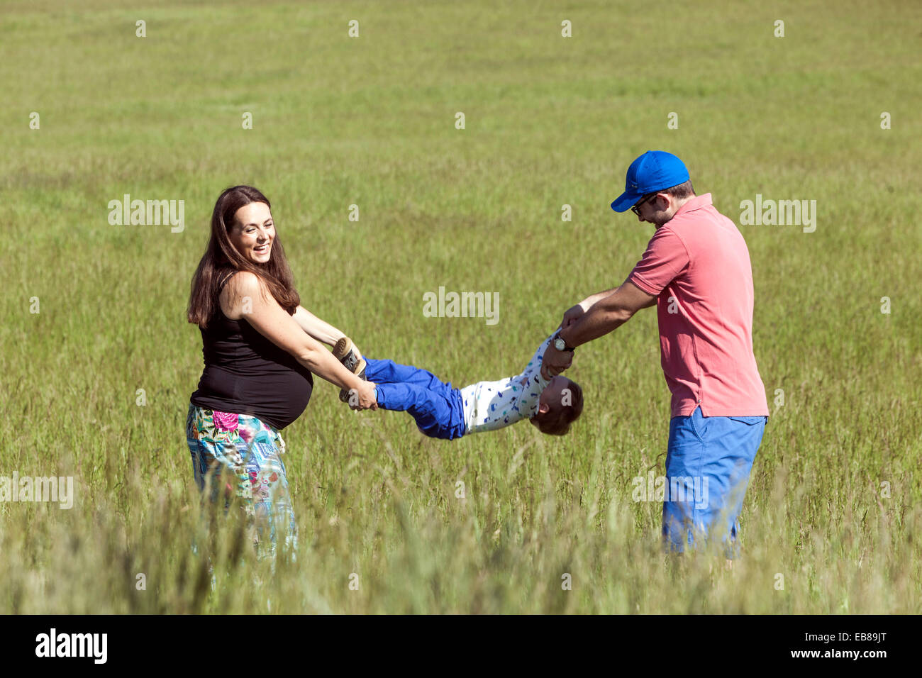Una familia feliz, una mujer embarazada con su marido y su niño a pasar tiempo juntos y jugar a juegos en una pradera de verano Foto de stock