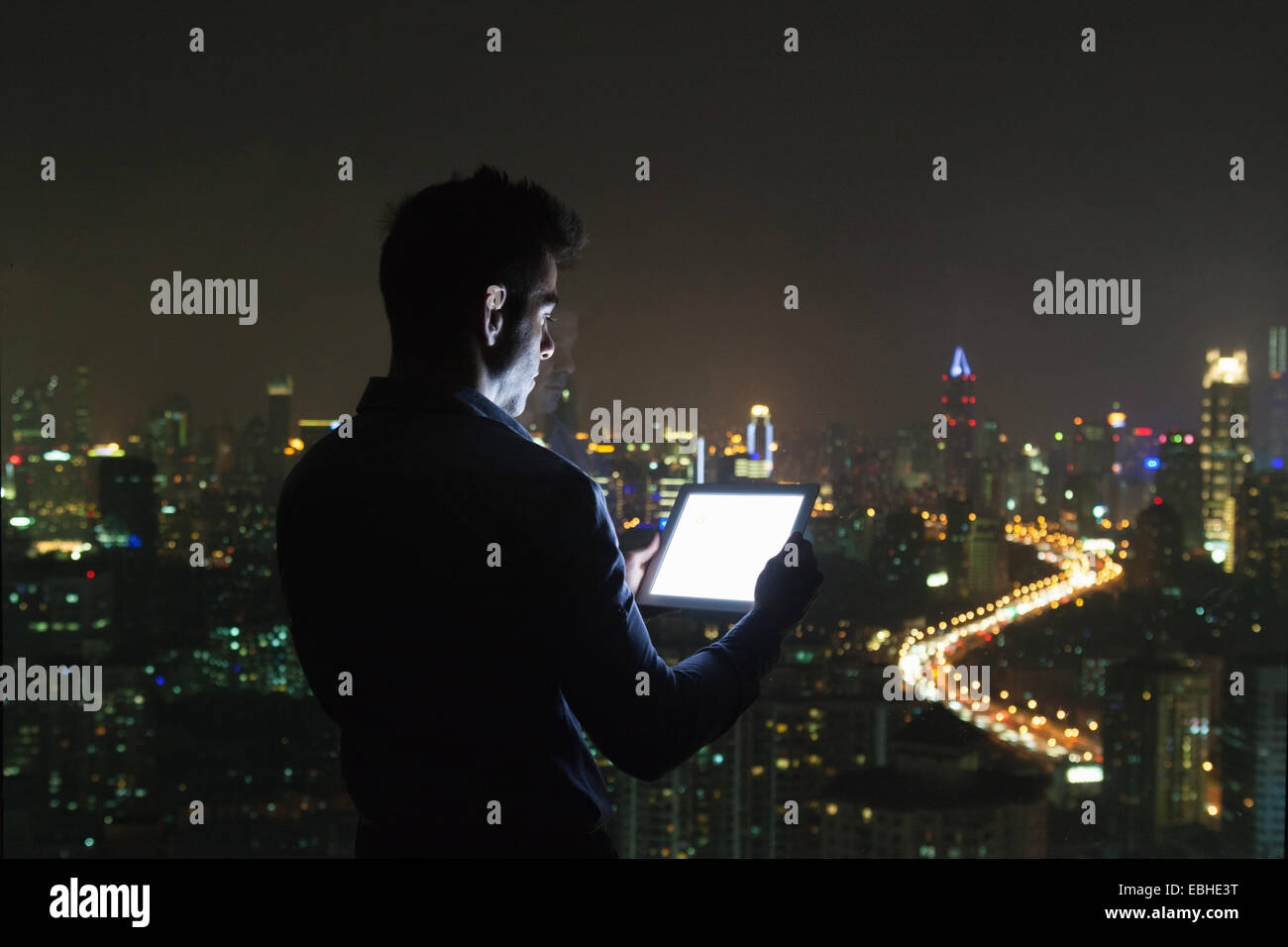 Siluetas de joven empresario mirando tableta digital delante de la ventana de la oficina de rascacielos en la noche, Shanghai, China Foto de stock