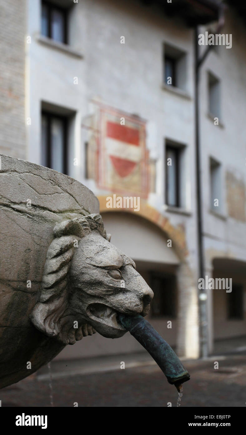 Fuente con cabeza de león en la parte delantera de un edificio medieval con fresco representando el escudo de Austria. Casco antiguo de la ciudad de Pordenone. Foto de stock