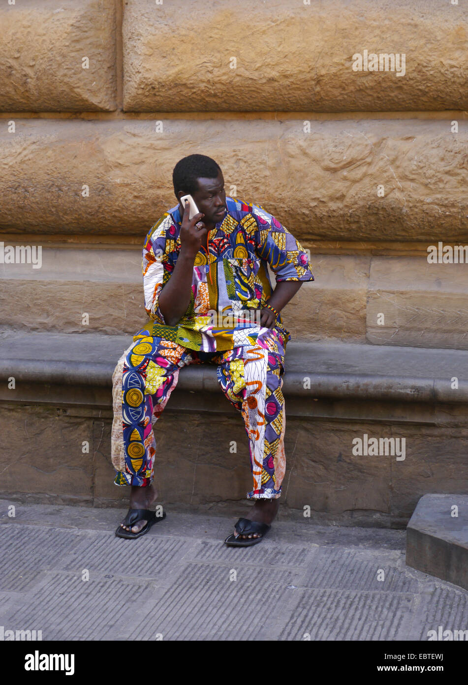 Un hombre afro-caribeño en un colorido traje de entramado sentado en un asiento de piedra en Florencia, Italia, hablando por un teléfono móvil Foto de stock
