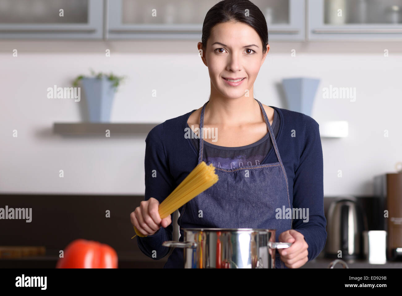 Joven ama de casa que prepara un saludable pasta italiana de pie en la estufa con un bote lleno de espaguetis con verduras frescas en th Foto de stock