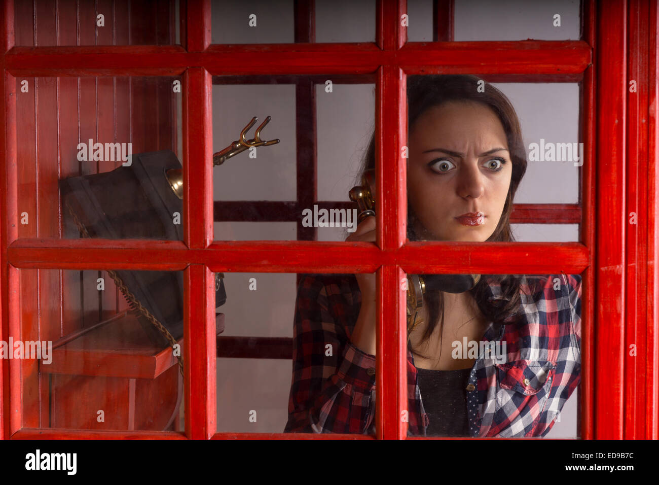 Mujer joven con una expresión horrorizada asomándose por la ventana de cristal del panel en una cabina telefónica pública como ella chats sobre un teléfono vintage. Foto de stock