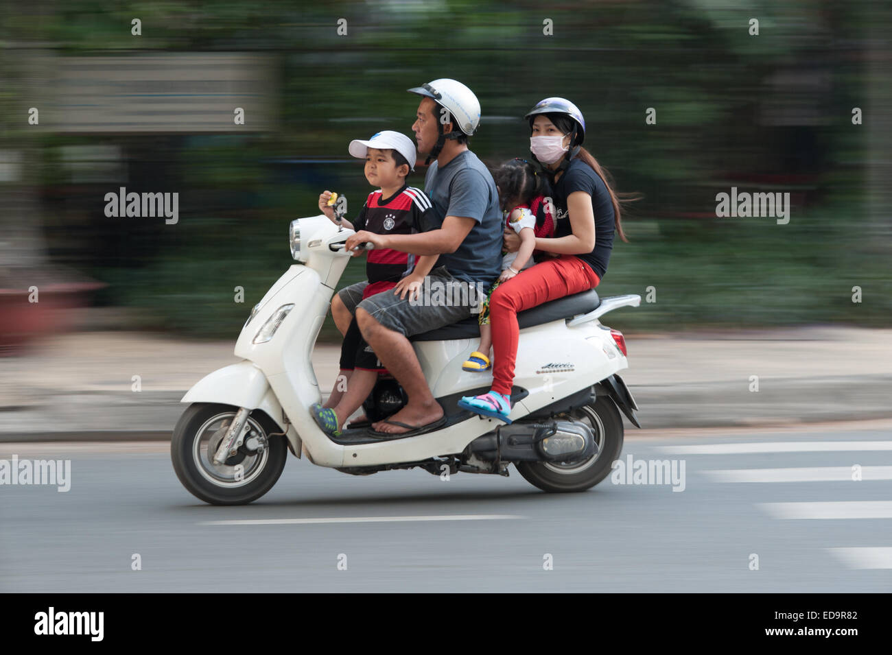 Familia de cuatro personas en moto scooter en Ciudad Ho Chi Minh, Vietnam dos niños comiendo no llevaban cascos Foto de stock
