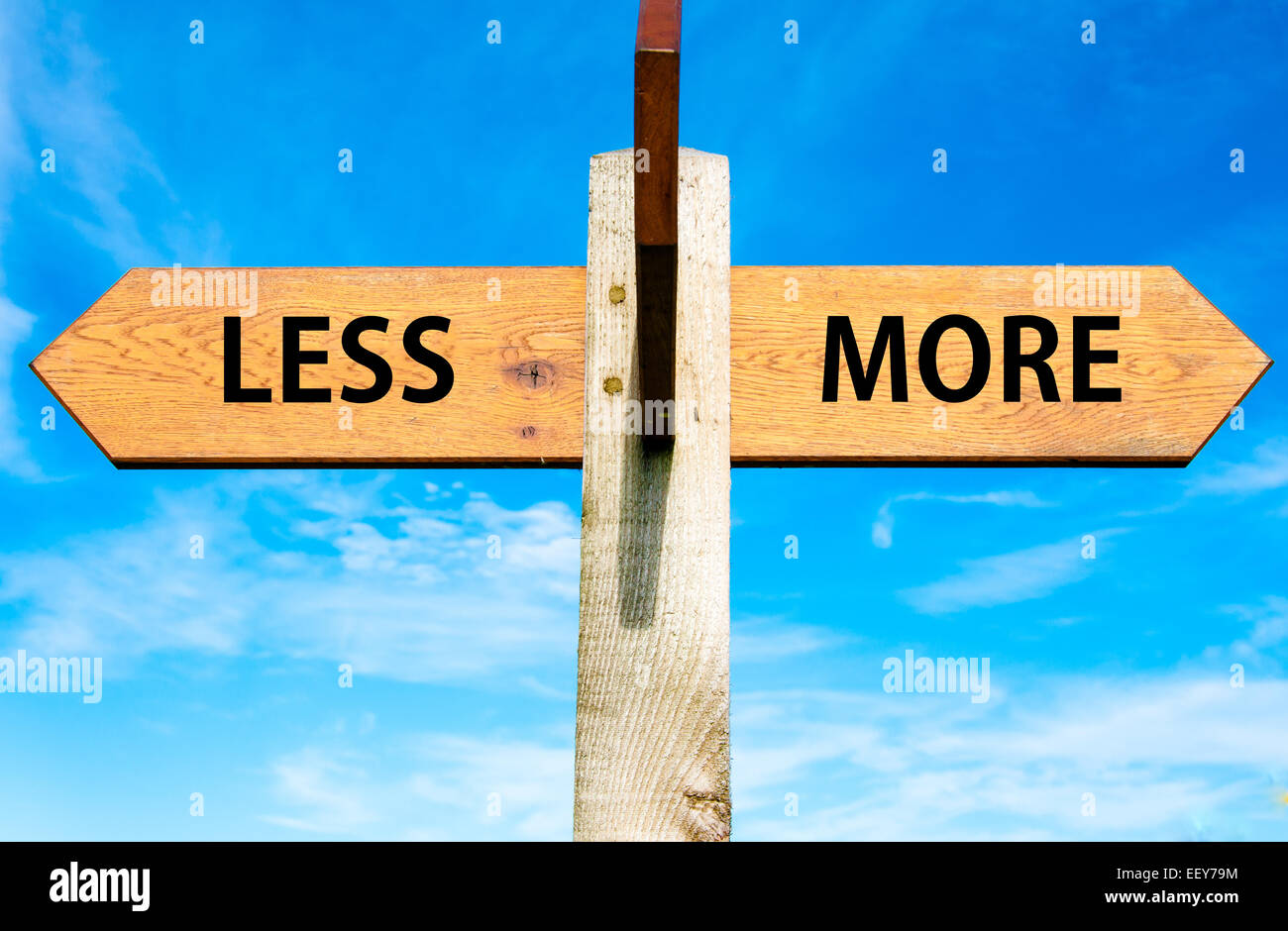 Cartel de madera con dos flechas opuestas sobre el azul claro del cielo, menos versus más mensajes Foto de stock